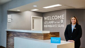 A reception desk with a 'Welcome to the Land of the Midnight Sun' sign, a 'Manager On Duty' sign, and a 'Dogs & Cats Allowed' sign. - Hampton Inn Anchorage (Anchorage)