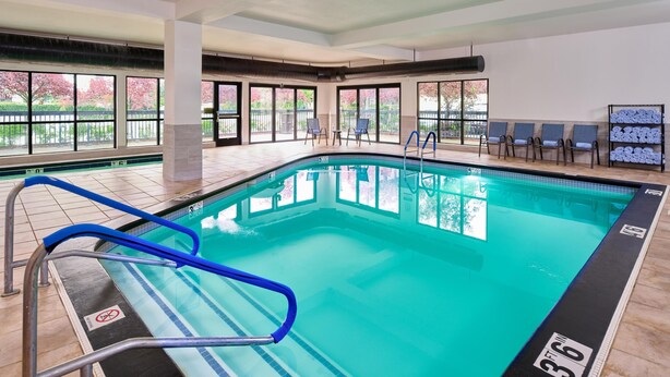 An indoor swimming pool with clear glass walls, blue poolside chairs, and a tiled floor. - Hampton Inn Anchorage (Anchorage)