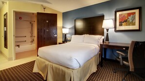 Quarto de hotel com cama, escrivaninha, cadeira, abajur e um quadro na parede.