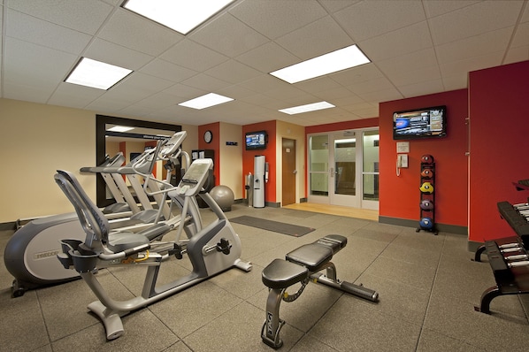 A gym with various exercise machines, a wall clock, and a television screen. - Hampton Inn Grand Rapids-South (Wyoming)