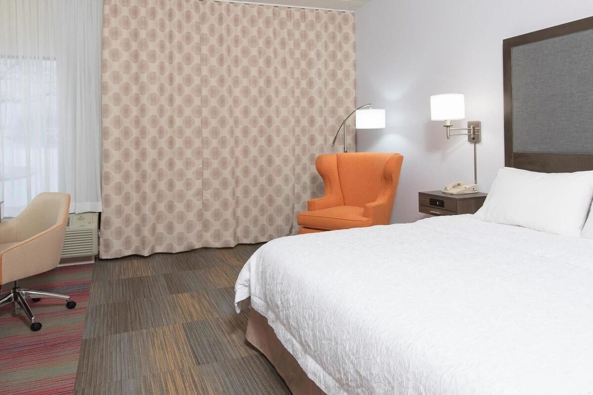 Ein Hotelzimmer mit einem Bett, einem orangefarbenen Sessel, einem Schreibtisch mit Lampe und einem Telefon.