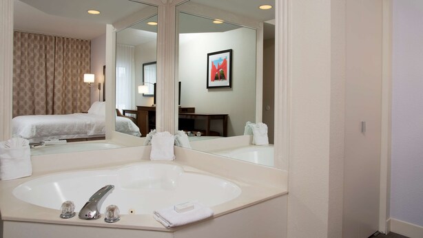 A hotel bathroom with a large mirror, a bathtub, and a bed visible in the reflection.
