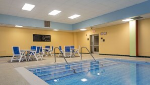 A swimming pool with blue water, surrounded by white and blue lounge chairs. There are signs on the wall and a door with an exit sign.