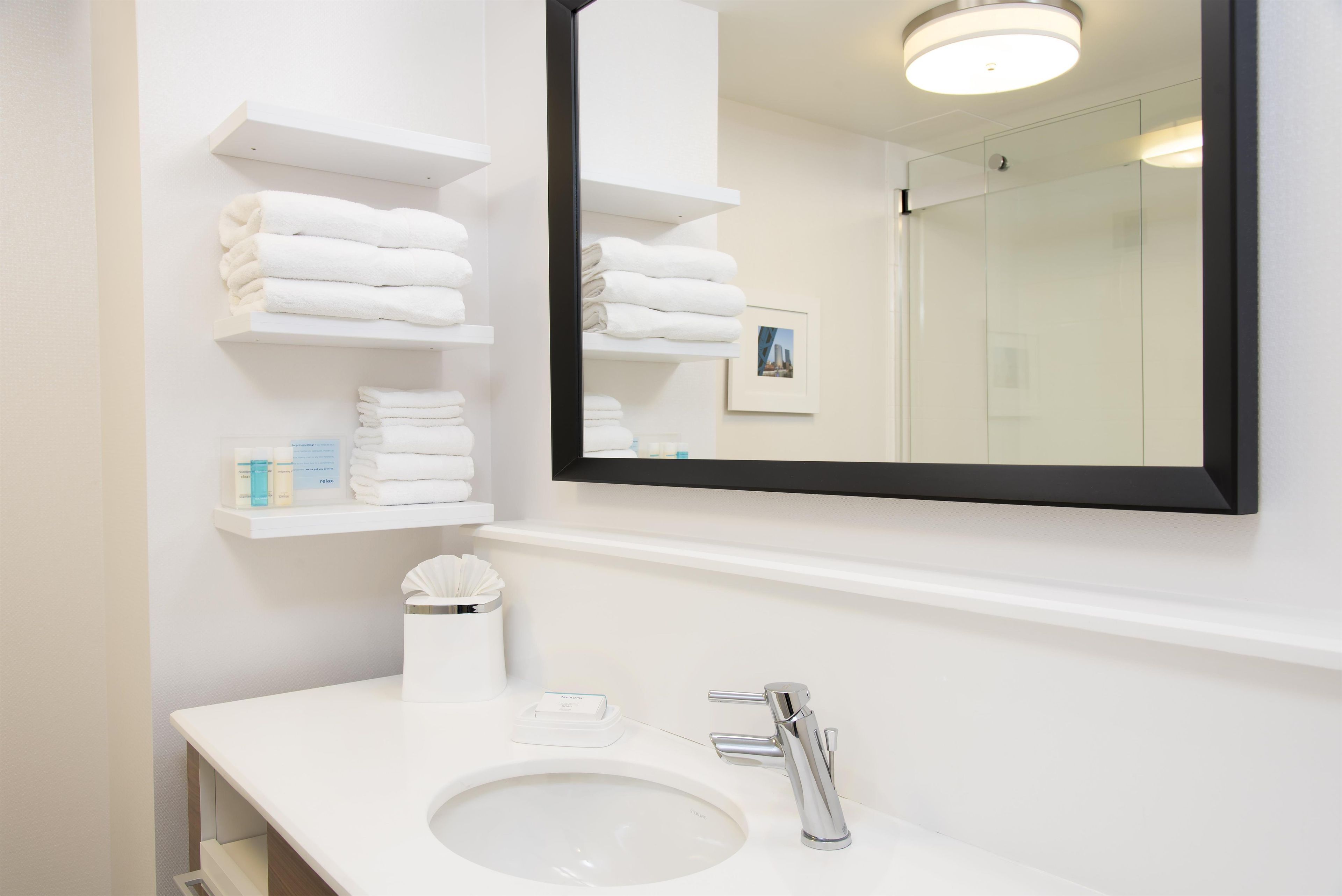 A modern bathroom with a white sink, chrome faucet, and a large mirror. There are white towel stacks on shelves and a small trash bin.