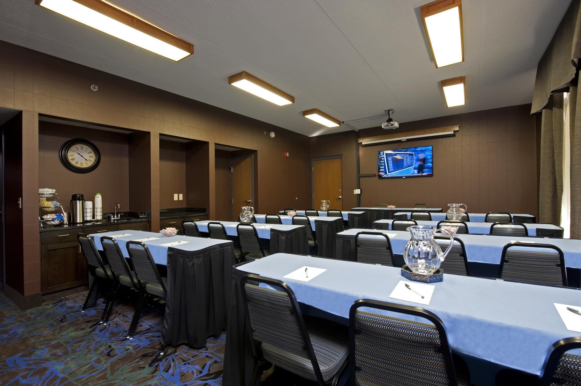 Un salón de conferencias con varias mesas, sillas, un reloj, una estación de café y una pantalla de televisión.