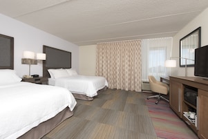 Quarto de hotel com duas camas, uma escrivaninha, uma cadeira, uma televisão e uma janela grande com cortinas.