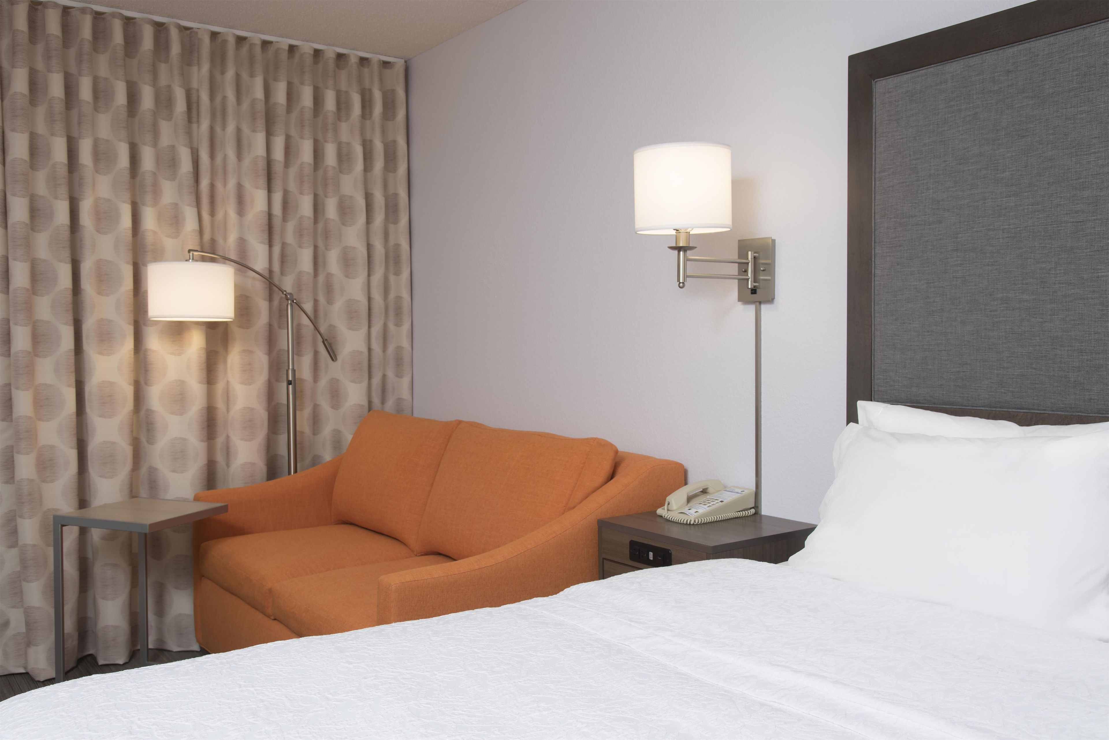 Une chambre d’hôtel avec un lit, un fauteuil orange, une table de chevet avec un téléphone et une lampe sur pied.