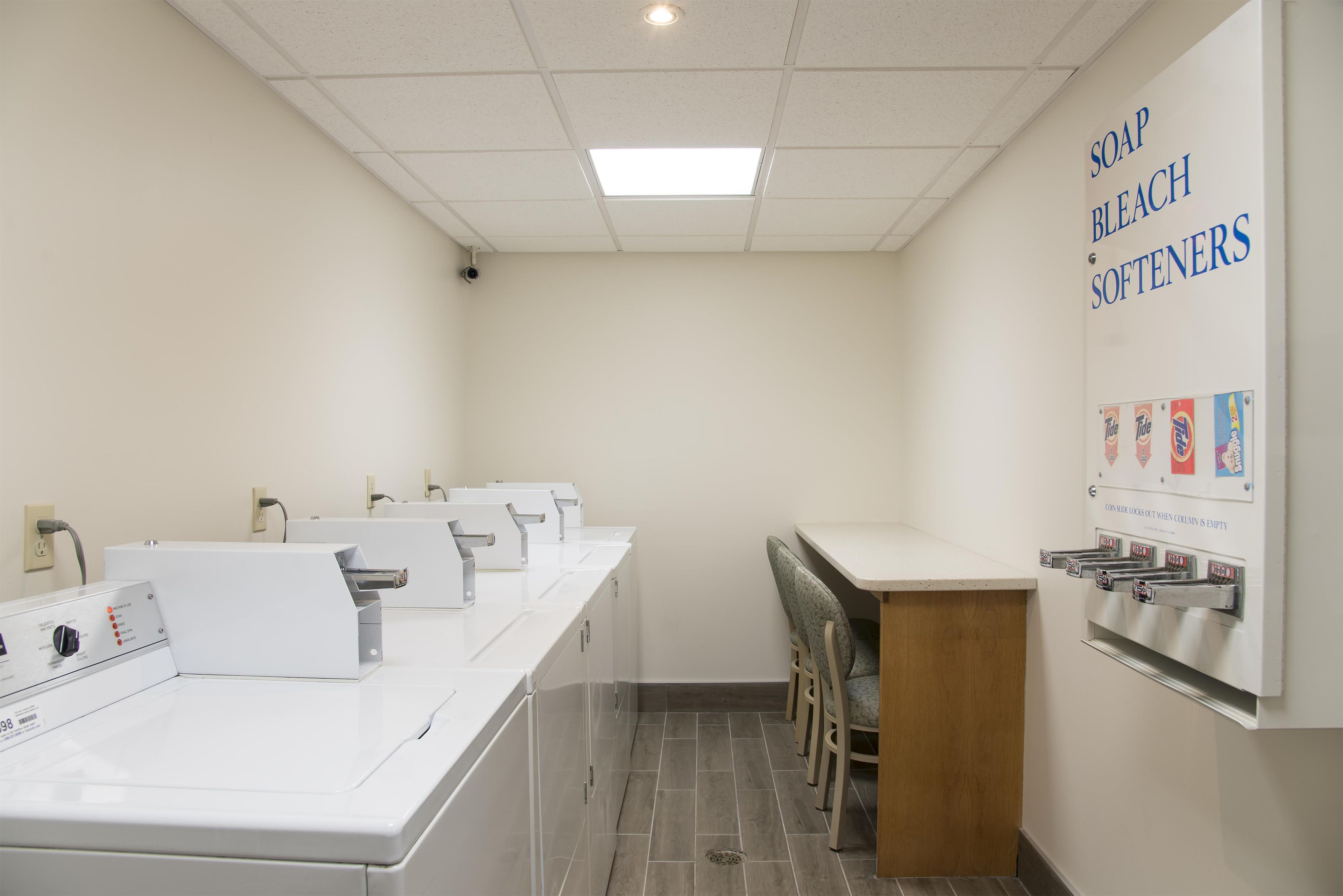 A laundromat with multiple washing machines, a seating area, and a wall with detergent brands.