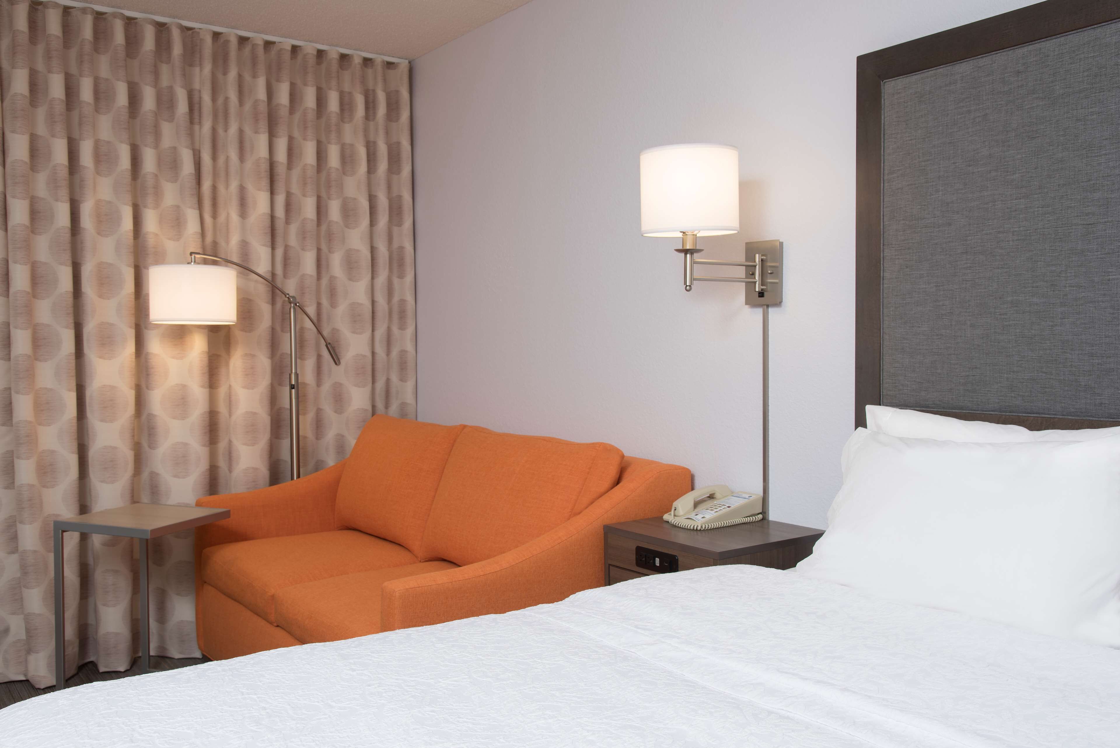 A hotel room with a bed, an orange armchair, a nightstand with a phone, and two wall-mounted lamps.