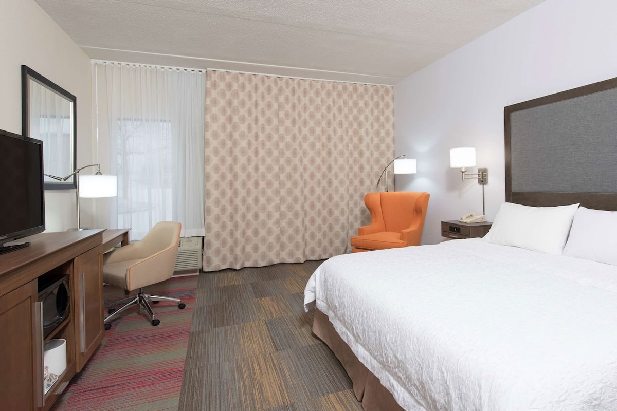 A hotel room with a bed, a desk with a TV, an orange chair, a nightstand, and a window with curtains.