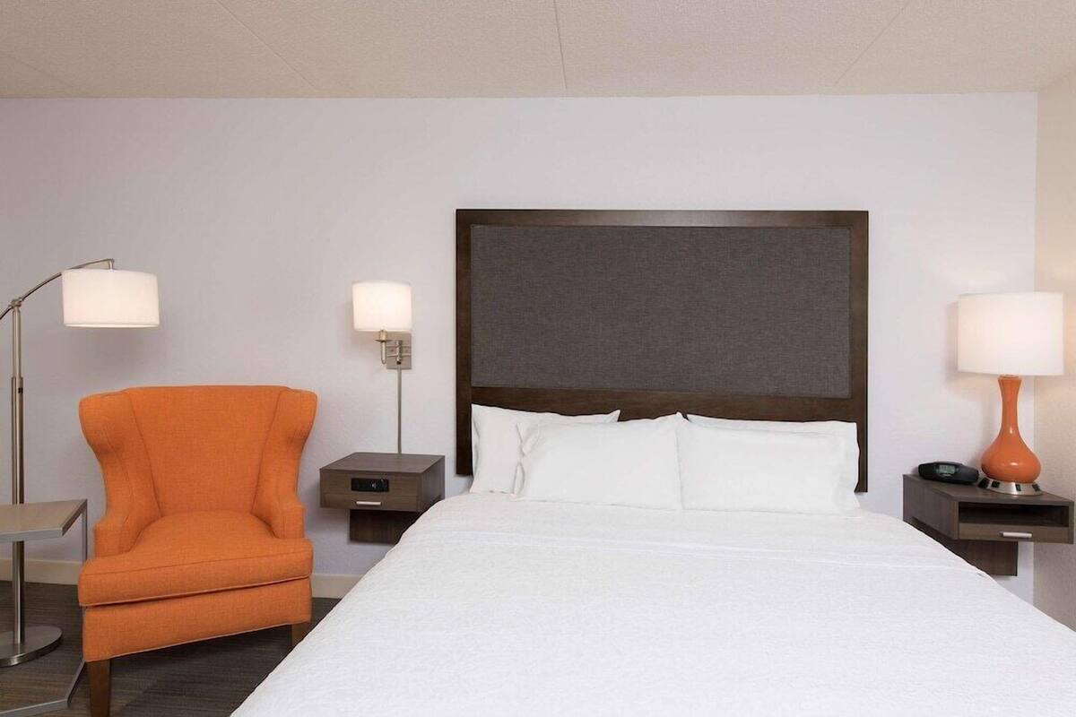 A hotel room with a large bed, an orange armchair, two bedside tables with lamps, and a modern headboard.