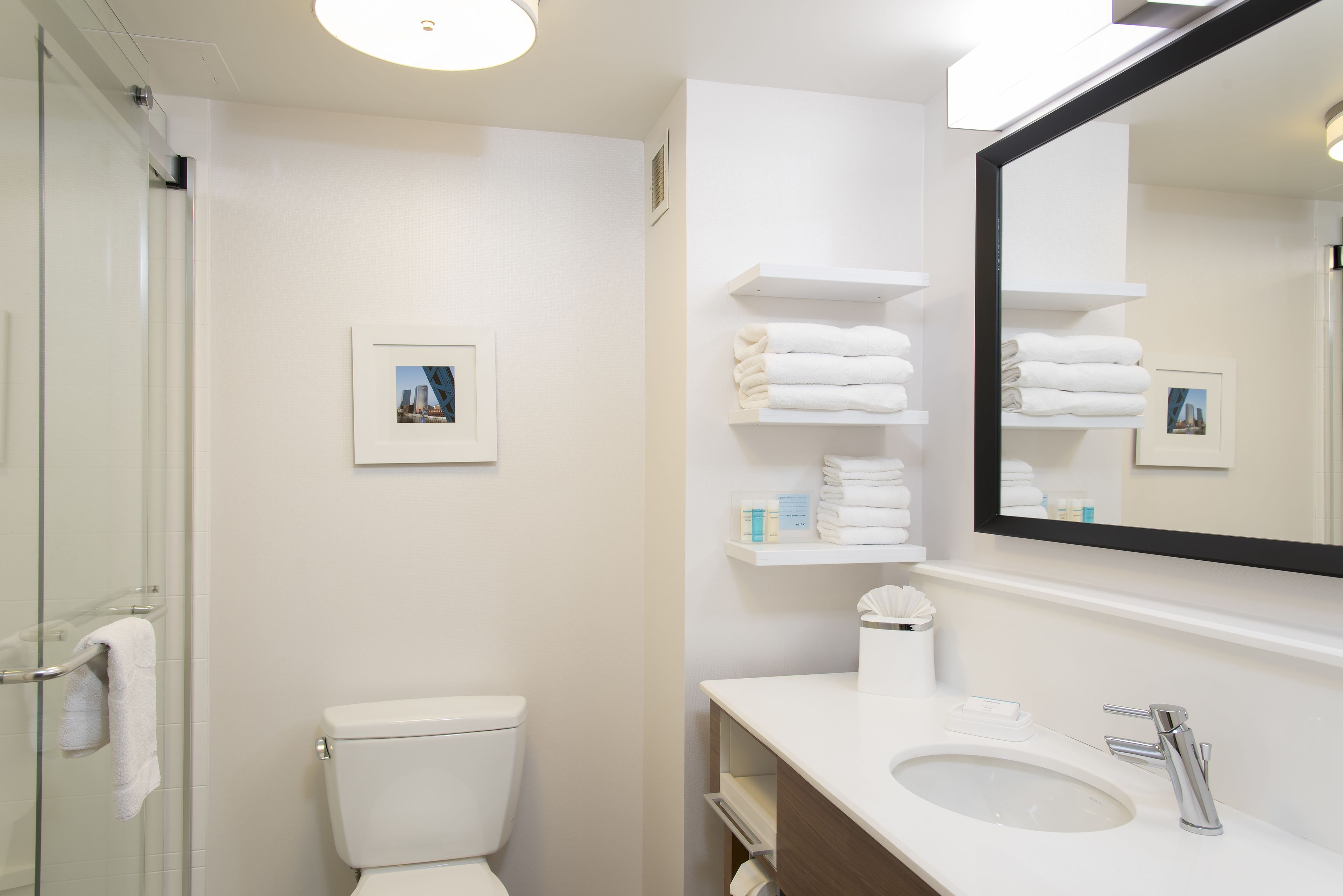 A modern bathroom with a glass shower enclosure, a white toilet, a sink with a mirror, and shelves with folded towels.