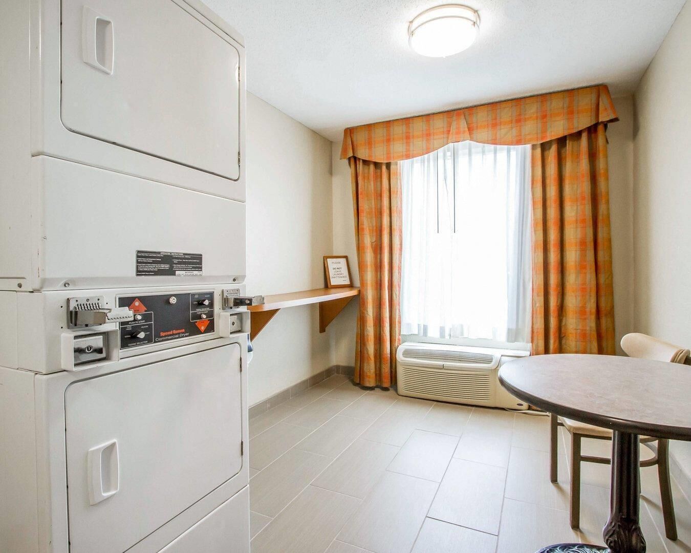 Een compacte hotelkamer met een kitchenette, een eethoek en een groot raam met gordijnen.