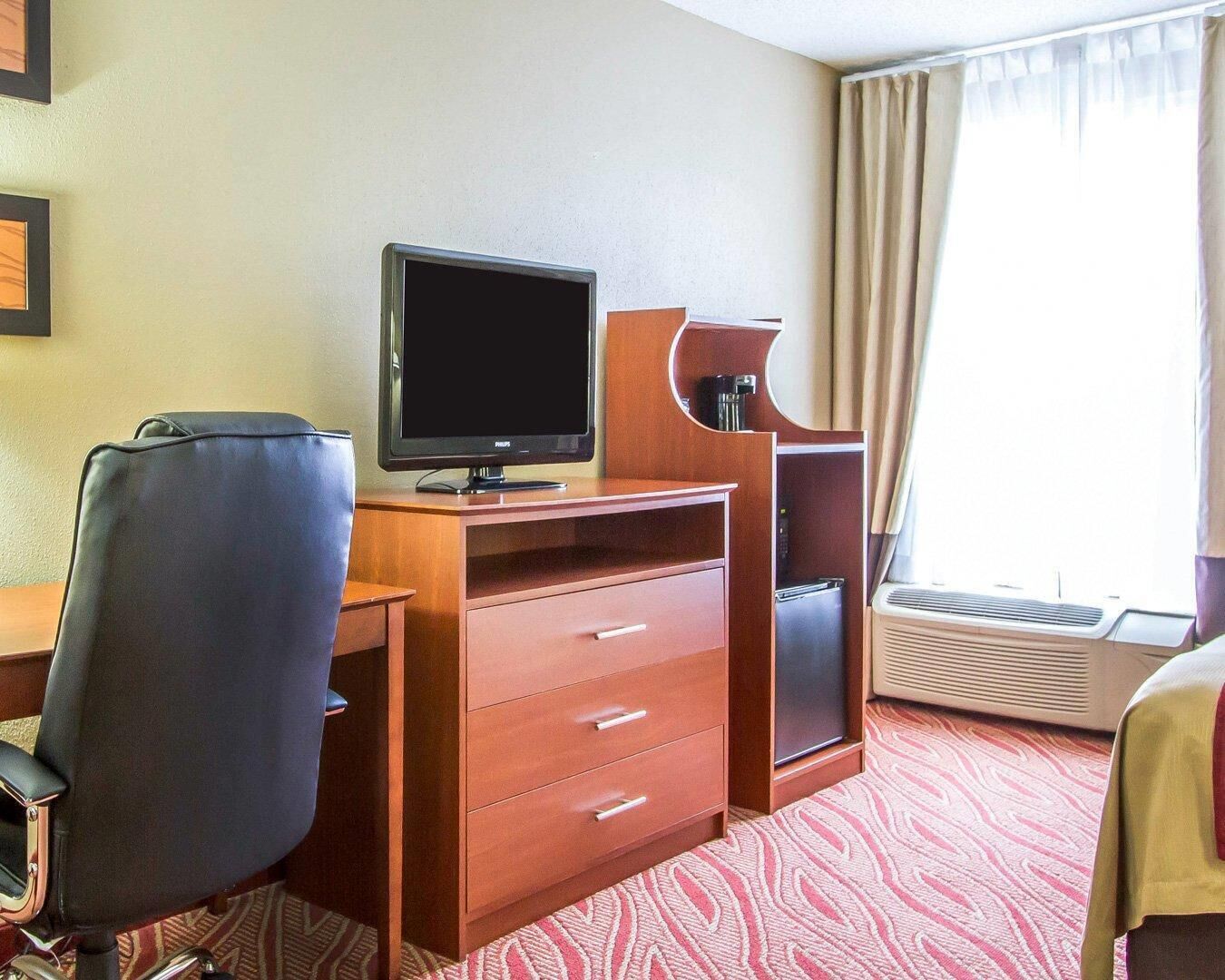 A hotel room with a wooden desk, a black office chair, a flat-screen TV, a coffee maker, a mini-fridge, and a window with curtains.