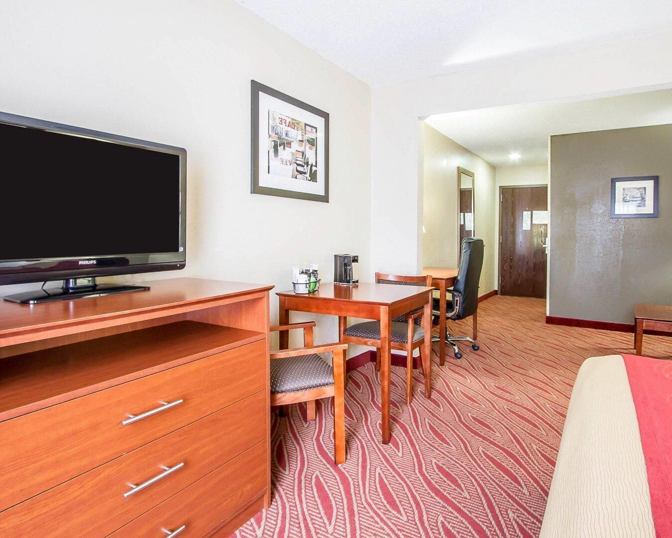 A hotel room with a flat-screen TV, wooden furniture, a patterned carpet, and a framed picture on the wall.
