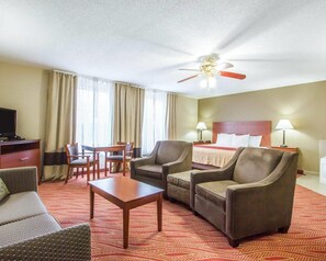 A hotel room with a bed, two armchairs, a sofa, a small table, a TV, a ceiling fan, and a window with curtains.