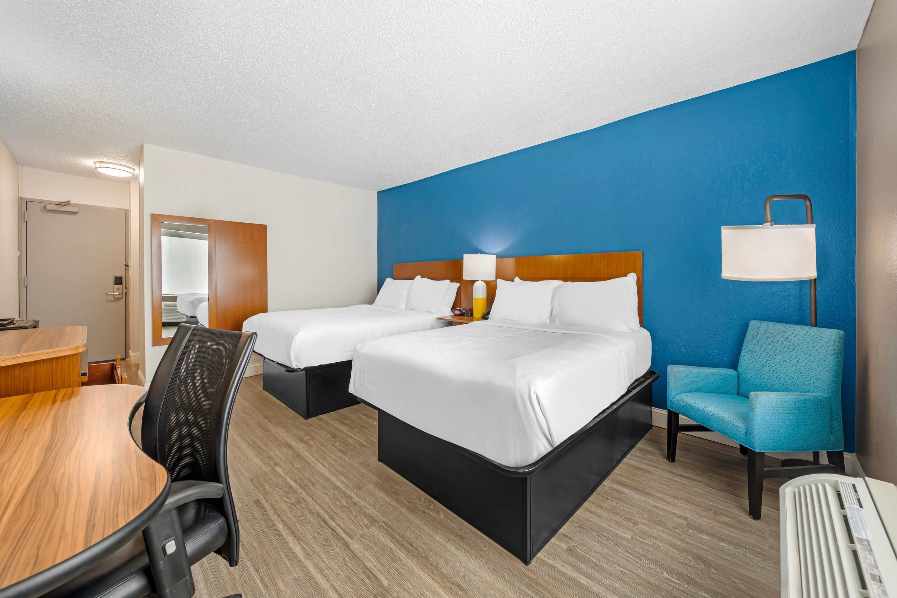 A hotel room with two beds, a desk, a chair, and a lamp.
