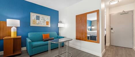 Ein Zimmer mit blauer Wand, einem türkisfarbenen Sofa, einem kleinen Tisch, einem Holznachttisch, einer Lampe, einem gerahmten Kunstwerk und einer zur Toilette führenden Tür.