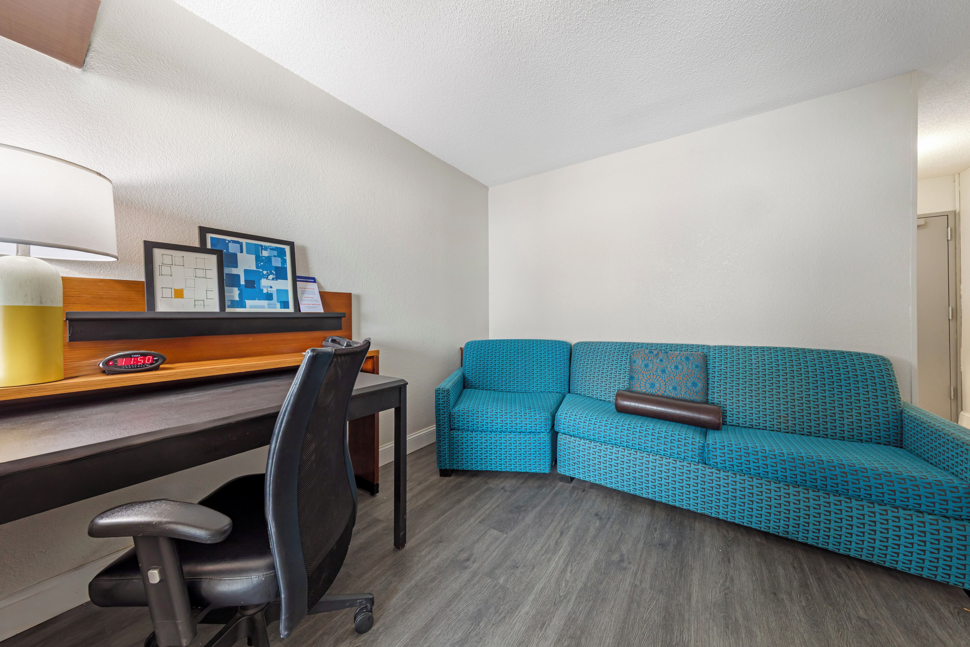 A room with a desk, a blue patterned sofa, a lamp, and framed pictures on the wall.