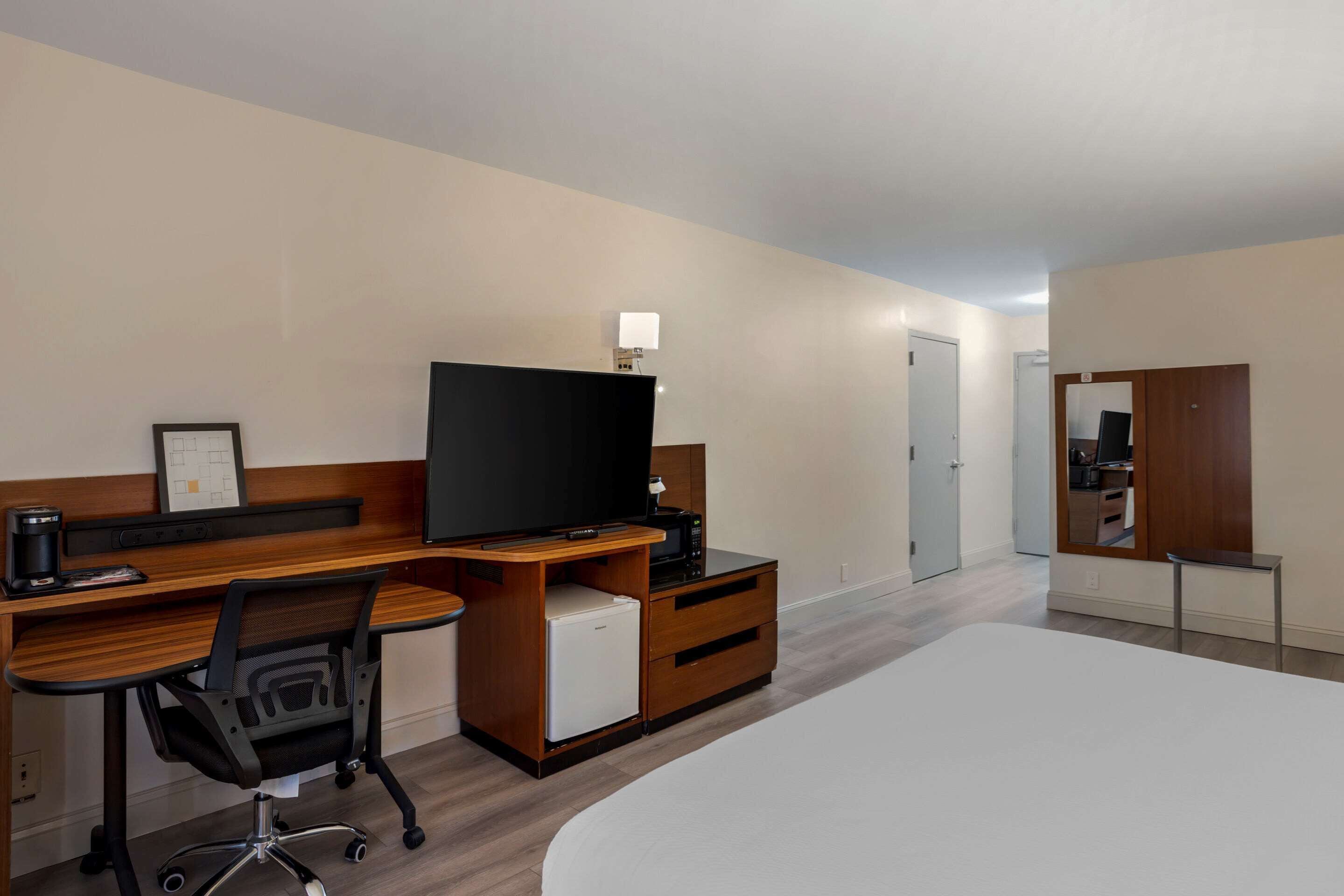 Una habitación de hotel con escritorio de madera, televisor, refrigerador pequeño, una silla, cama y espejo.