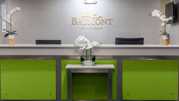 Un comptoir d’accueil avec une enseigne Baymont by Wyndham, une petite table, deux orchidées en pot et un mur d’accent vert.