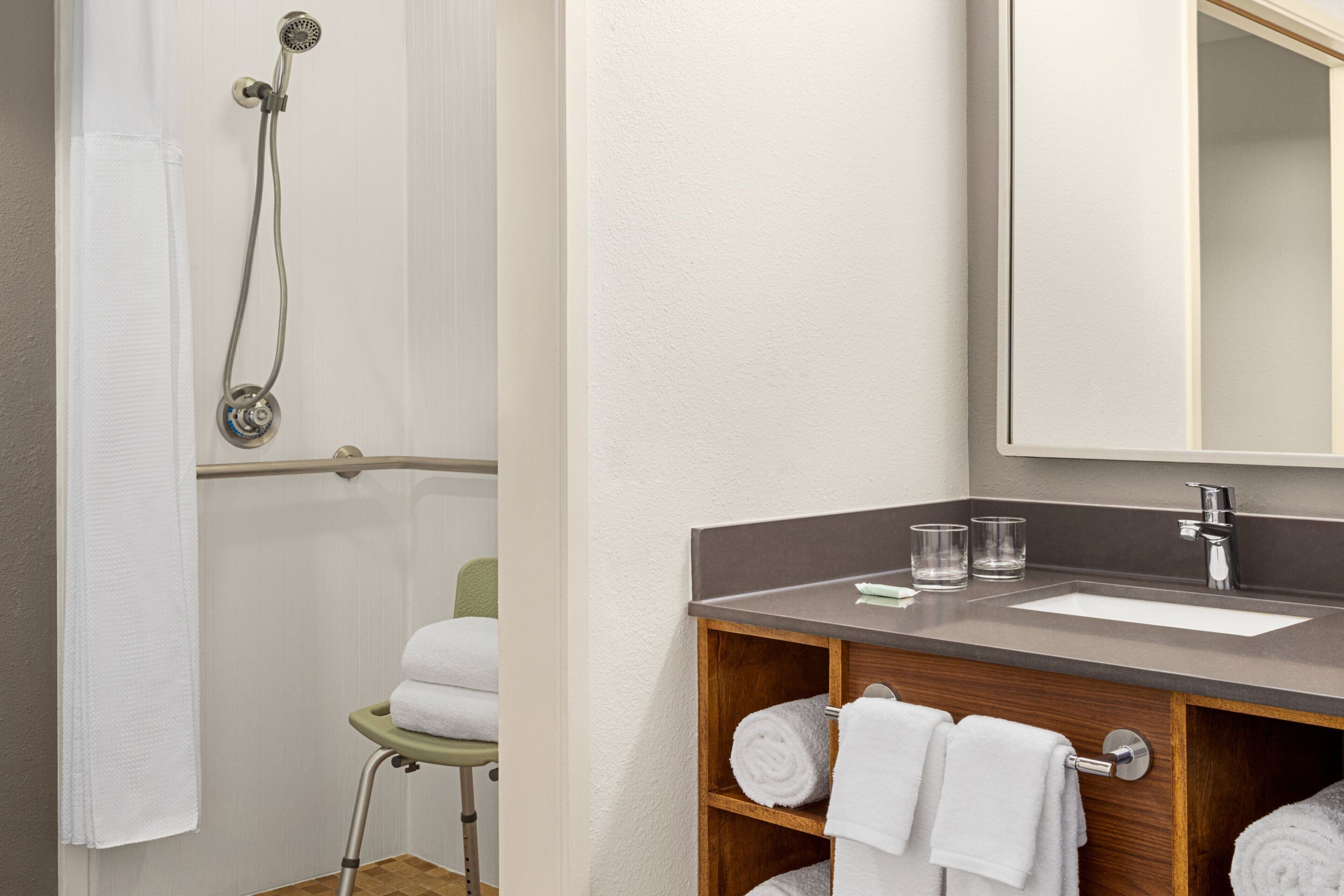 A bathroom with a shower, a sink, and a mirror. There are towels and glasses on the counter.