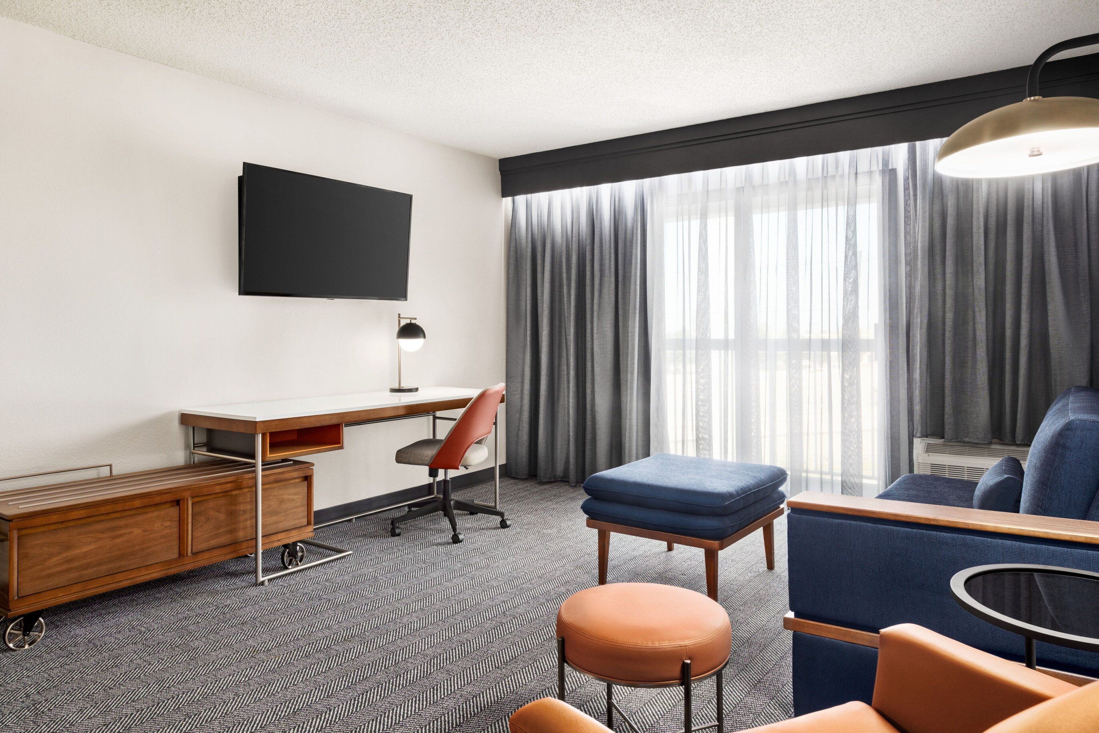 A modern hotel room with a flat-screen TV mounted on the wall, a wooden desk with a chair, a blue sofa, an orange ottoman, and a round table.