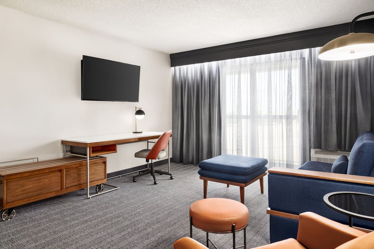 A modern hotel room with a flat-screen TV mounted on the wall, a wooden desk with a chair, a blue sofa, an orange ottoman, and a round table.