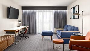 Ein modernes Hotelzimmer mit Flachbildfernseher, Schreibtisch, blauem Sofa, orangefarbenem Sessel und einem großen Fenster mit Vorhängen.