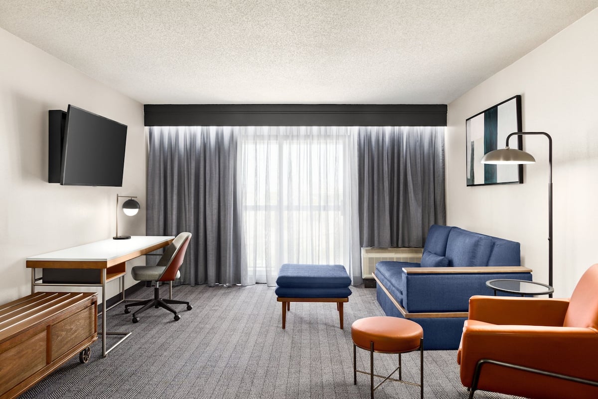A modern hotel room with a flat-screen TV, a desk, a blue sofa, an orange armchair, and a large window with curtains.