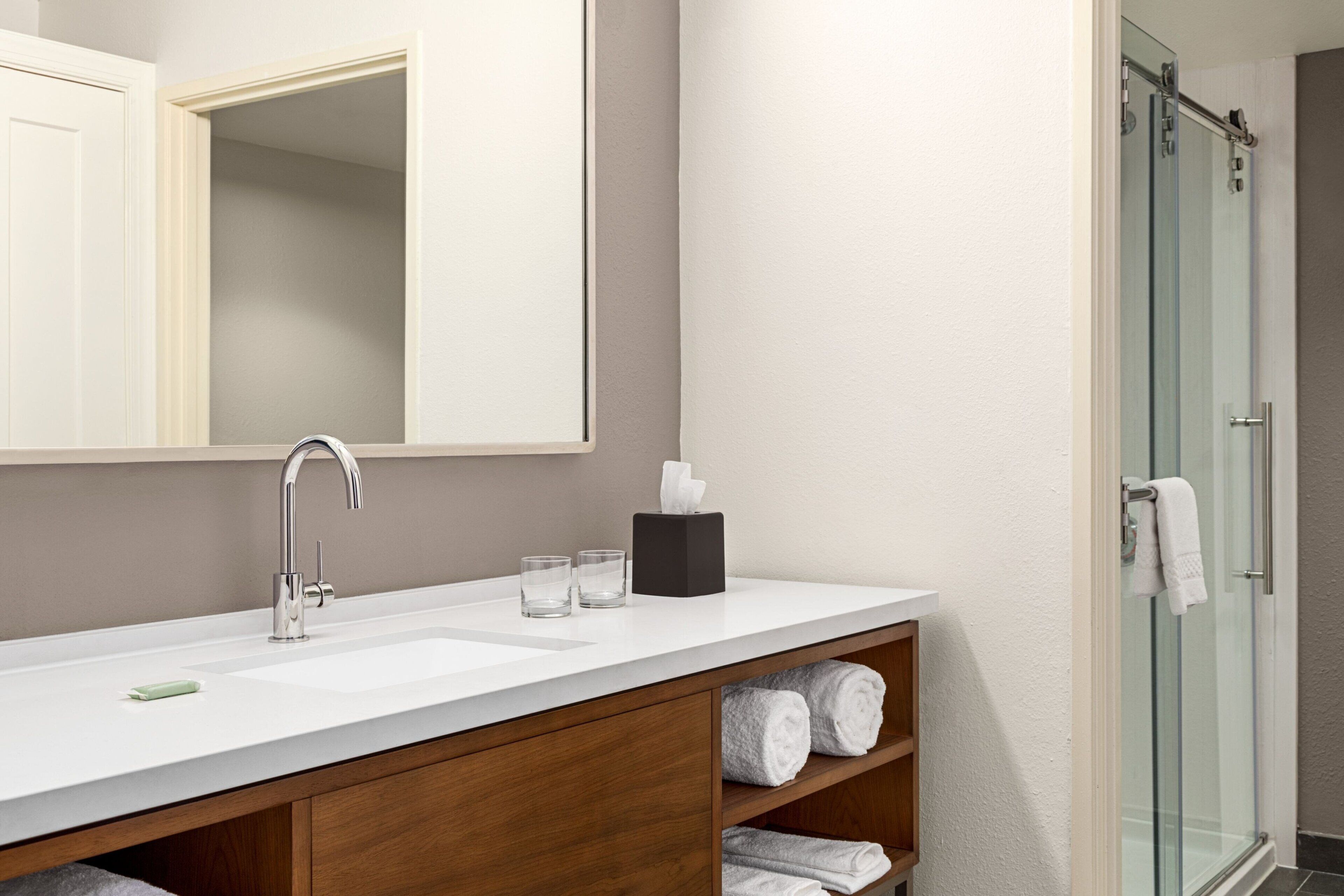 A modern bathroom with a large mirror, a white sink, a glass shower enclosure, and wooden shelves with towels.
