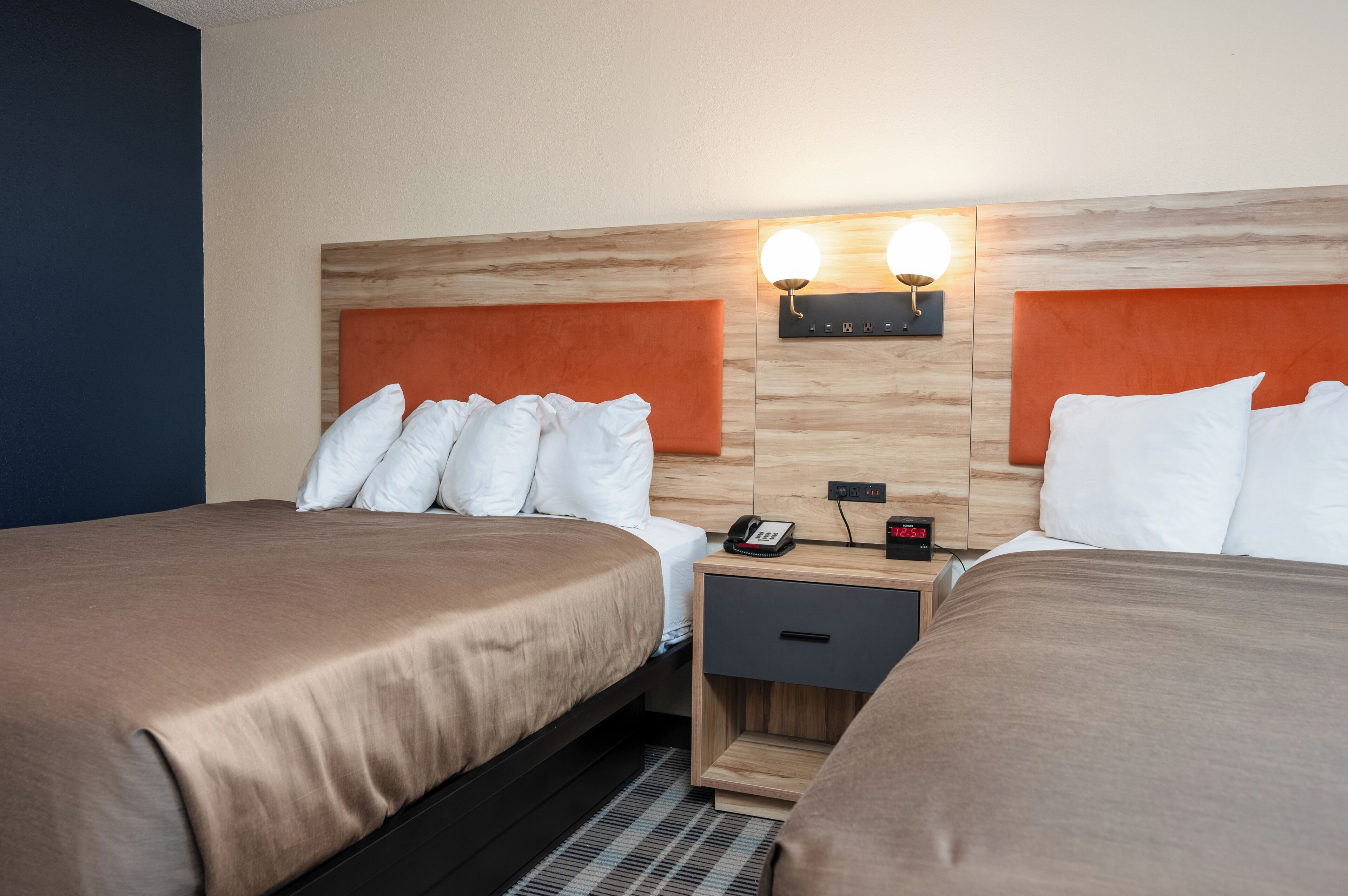 Une chambre d’hôtel avec deux lits, une tête de lit en bois, une table de chevet avec un téléphone et une horloge, et des lampes fixées au mur.