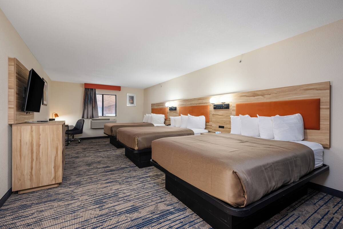 Quarto de hotel com quatro camas, uma escrivaninha, televisão e janela com cortinas.