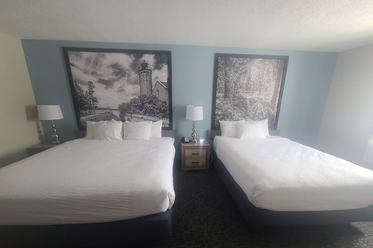 Quarto de hotel com duas camas, cada uma com lençóis e almofadas brancas, uma mesa de cabeceira com abajur e duas fotografias a preto e branco emolduradas na parede.