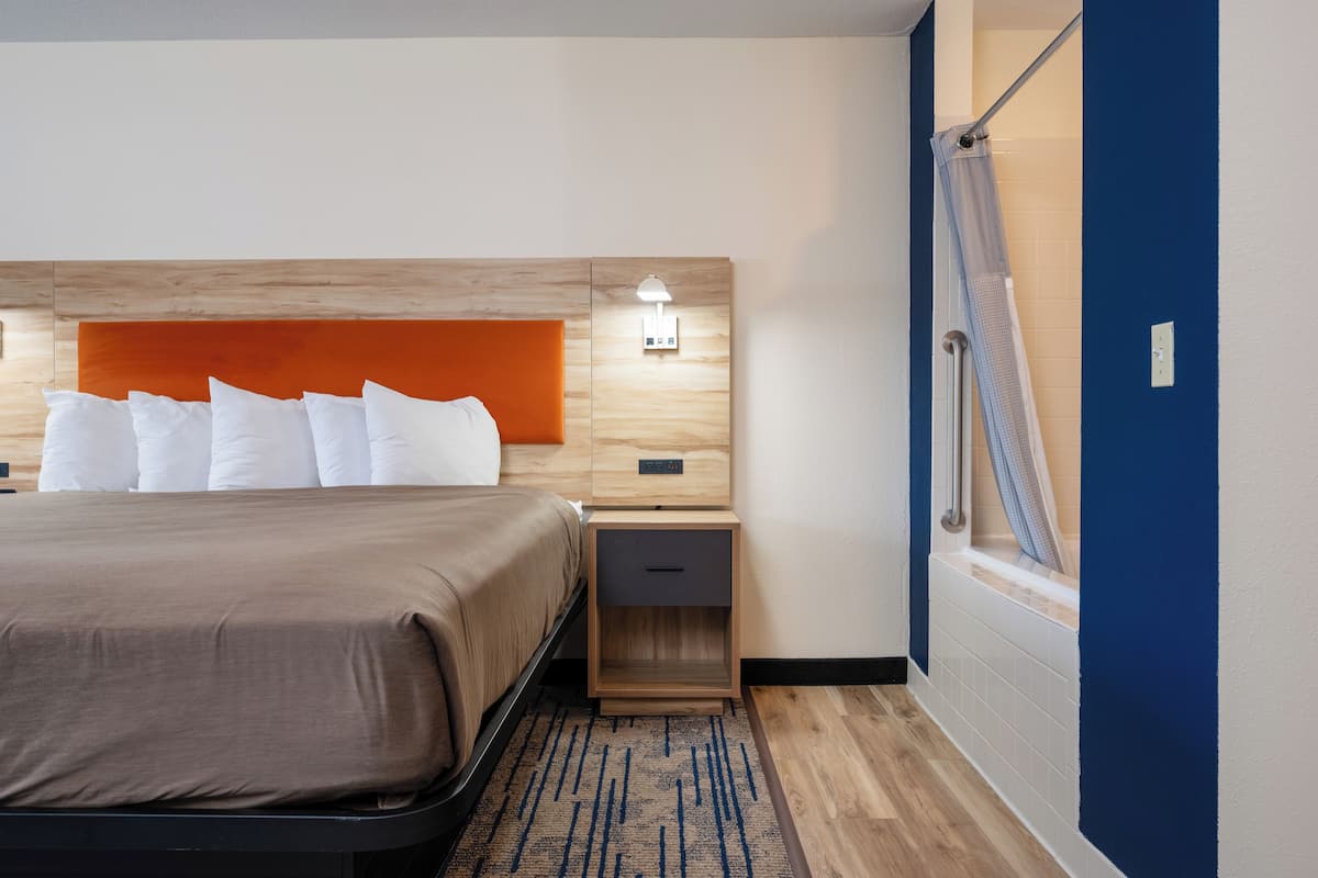 Quarto de hotel com cama, mesa de cabeceira, luminária na parede, área de chuveiro e cabeceira de madeira.