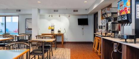 A modern hotel room with a dining area, a coffee bar, and a flat-screen TV mounted on the wall.