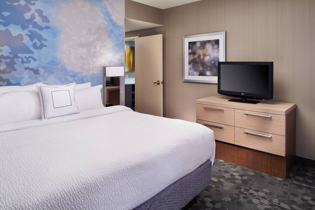 A hotel room with a large bed, a television on a wooden dresser, a door leading to a bathroom, and a wall with a camouflage pattern.