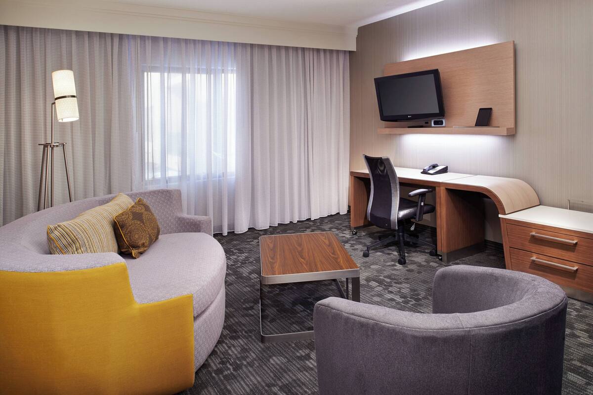 A hotel room with a sofa, armchair, desk, and a television mounted on the wall.