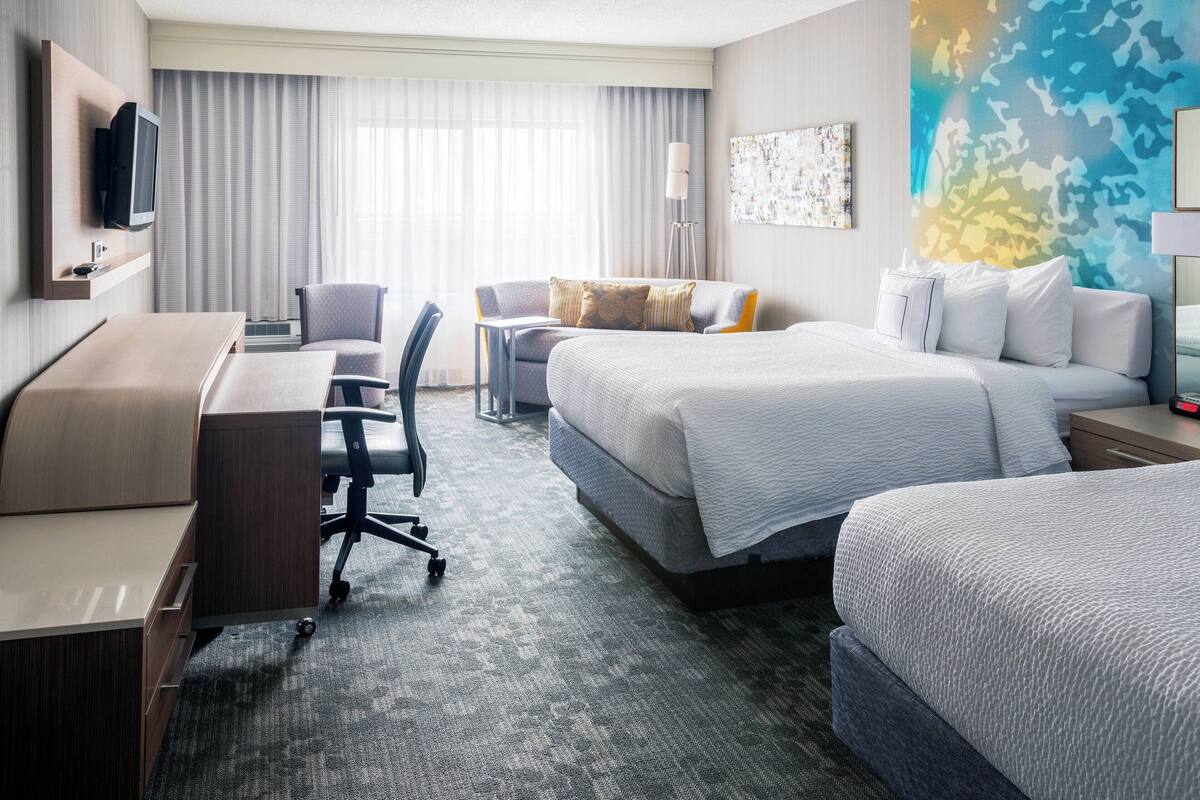 A hotel room with a desk, two beds, a chair, a television, and a large window with curtains.