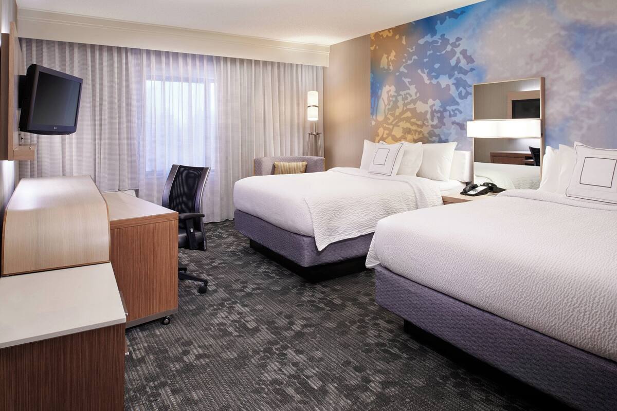 A hotel room with two beds, a desk, a chair, a TV, and a large window with curtains.