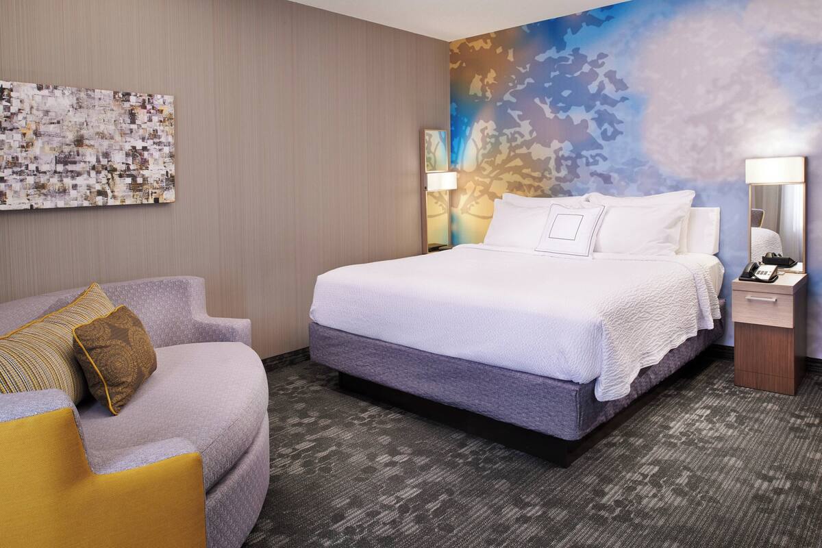 A hotel room with a large bed, a sofa, a wall art piece, and a nightstand with a lamp.