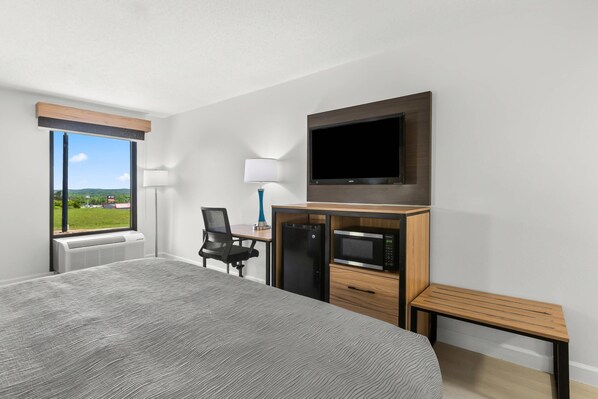 A hotel room with a large bed, a desk with a computer, a flat-screen TV, a microwave, and a window with a view of greenery.