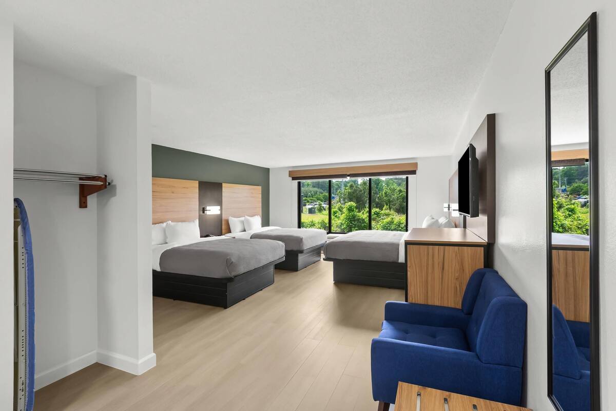 A modern hotel room with three beds, a blue armchair, a wooden desk, and a large window offering a view of greenery.