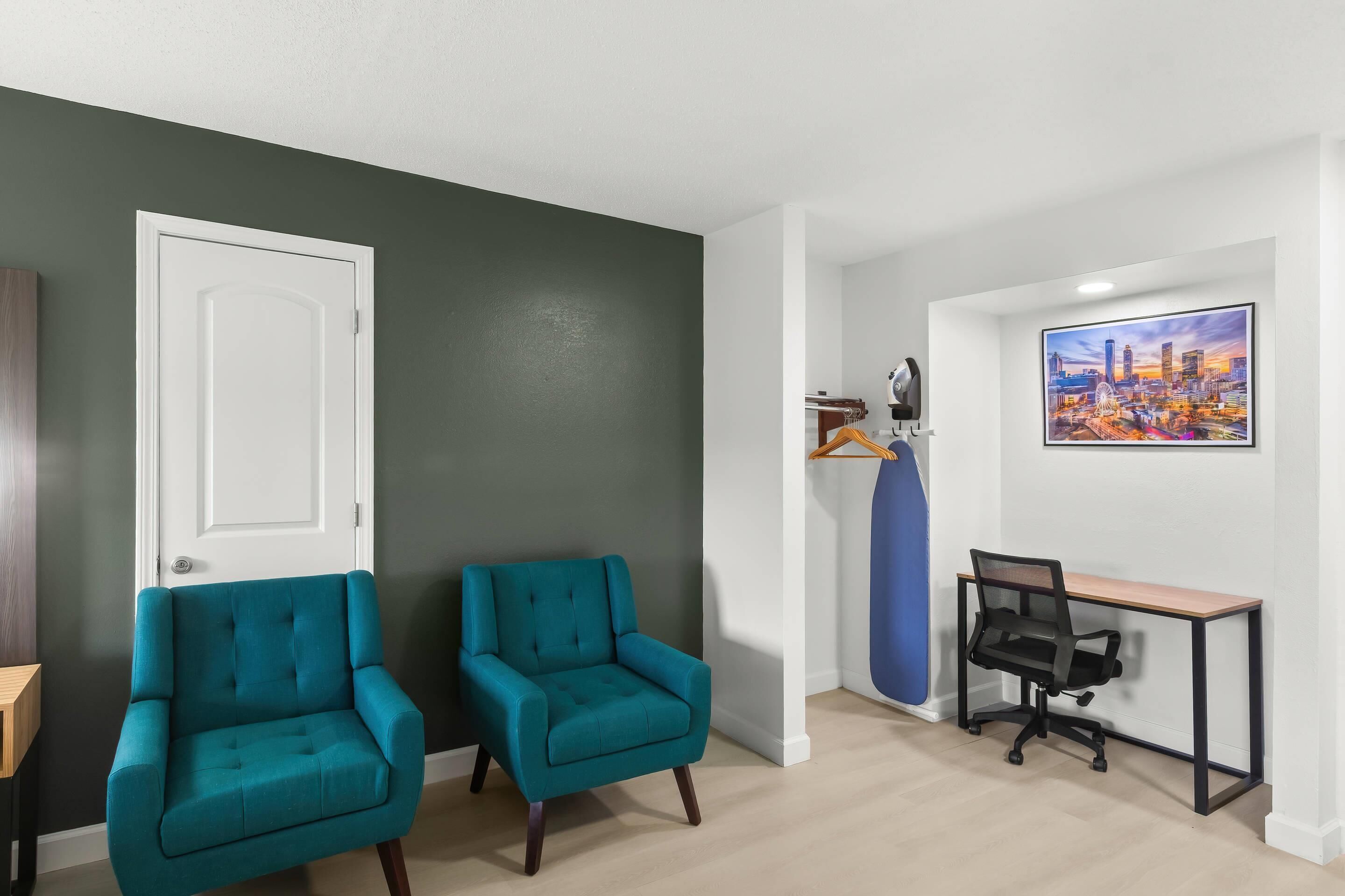 A room with two teal armchairs, a black office chair, a desk, and a cityscape painting on the wall.
