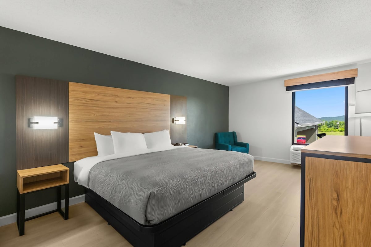 A modern bedroom with a large bed, a wooden headboard, a nightstand, a teal chair, and a window with a view.