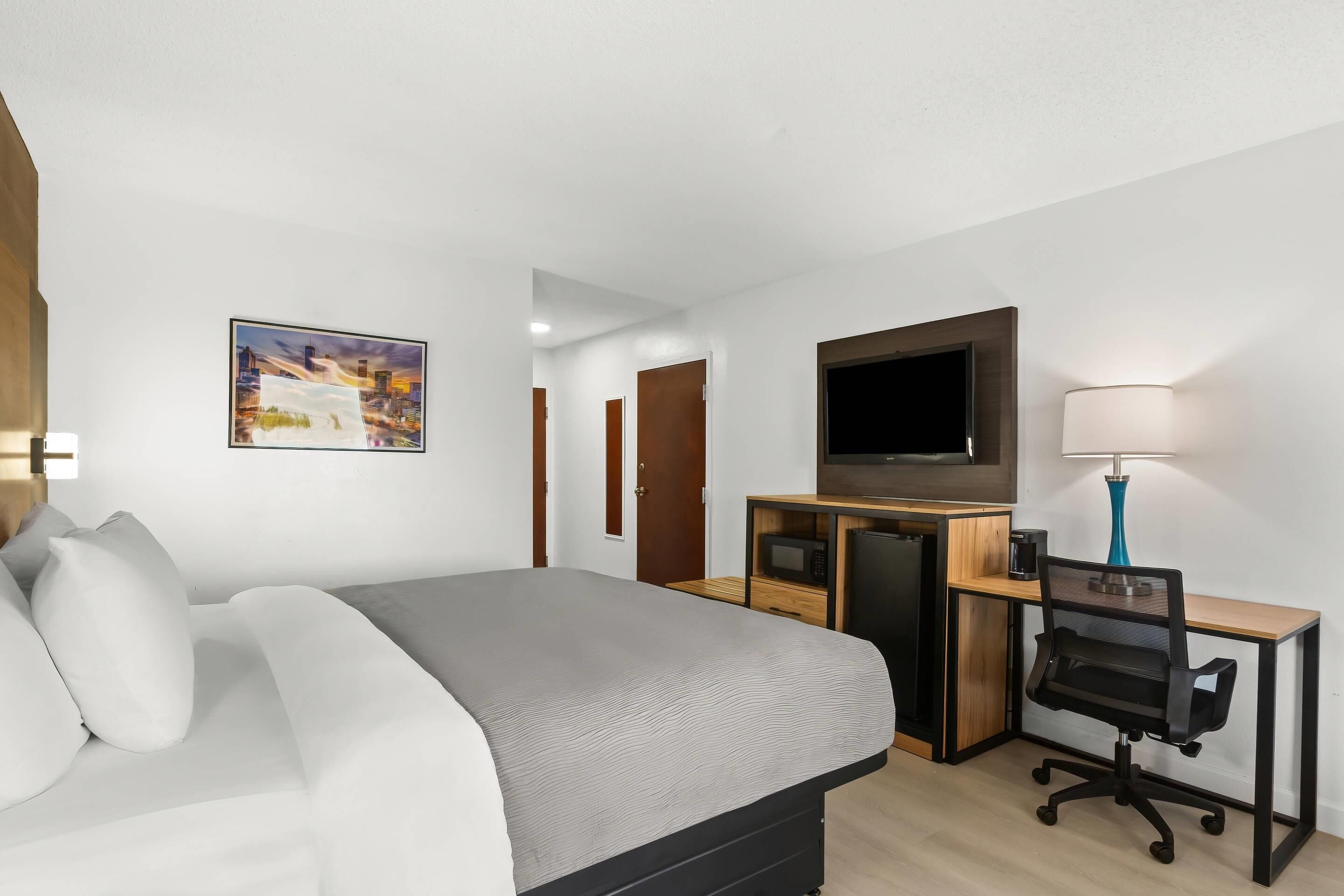 A hotel room with a bed, desk, chair, TV, and a painting on the wall.