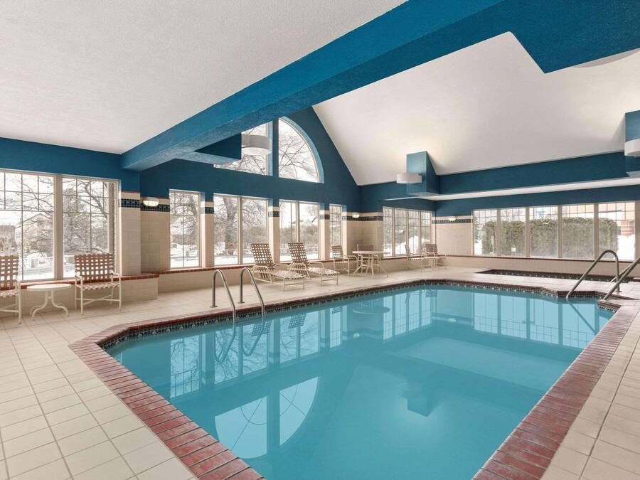An indoor swimming pool with clear blue water, surrounded by white chairs and tables, large windows, and a high ceiling with blue beams.