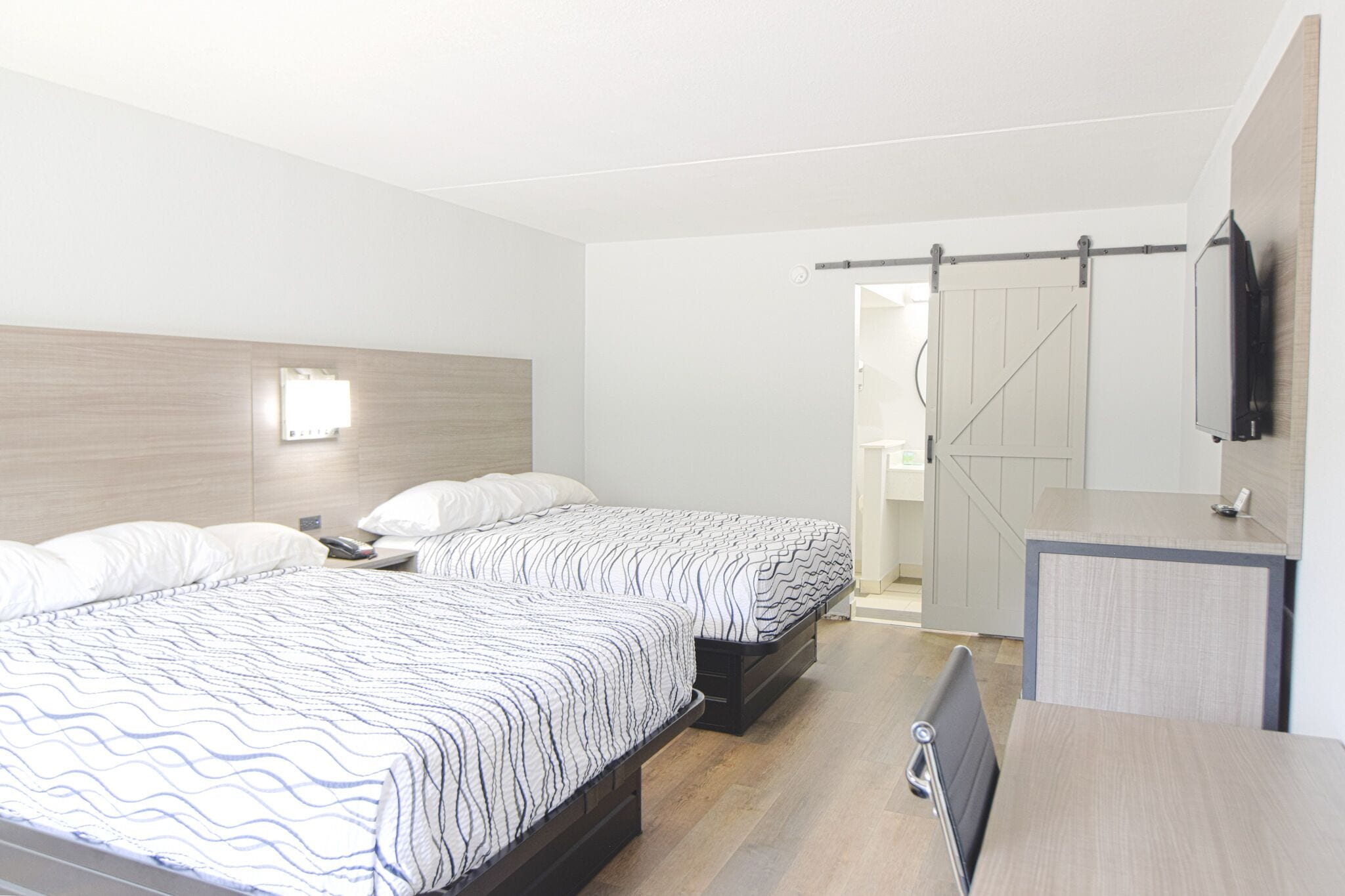 A modern hotel room with two beds, a desk, a TV, and a sliding door to a bathroom.
