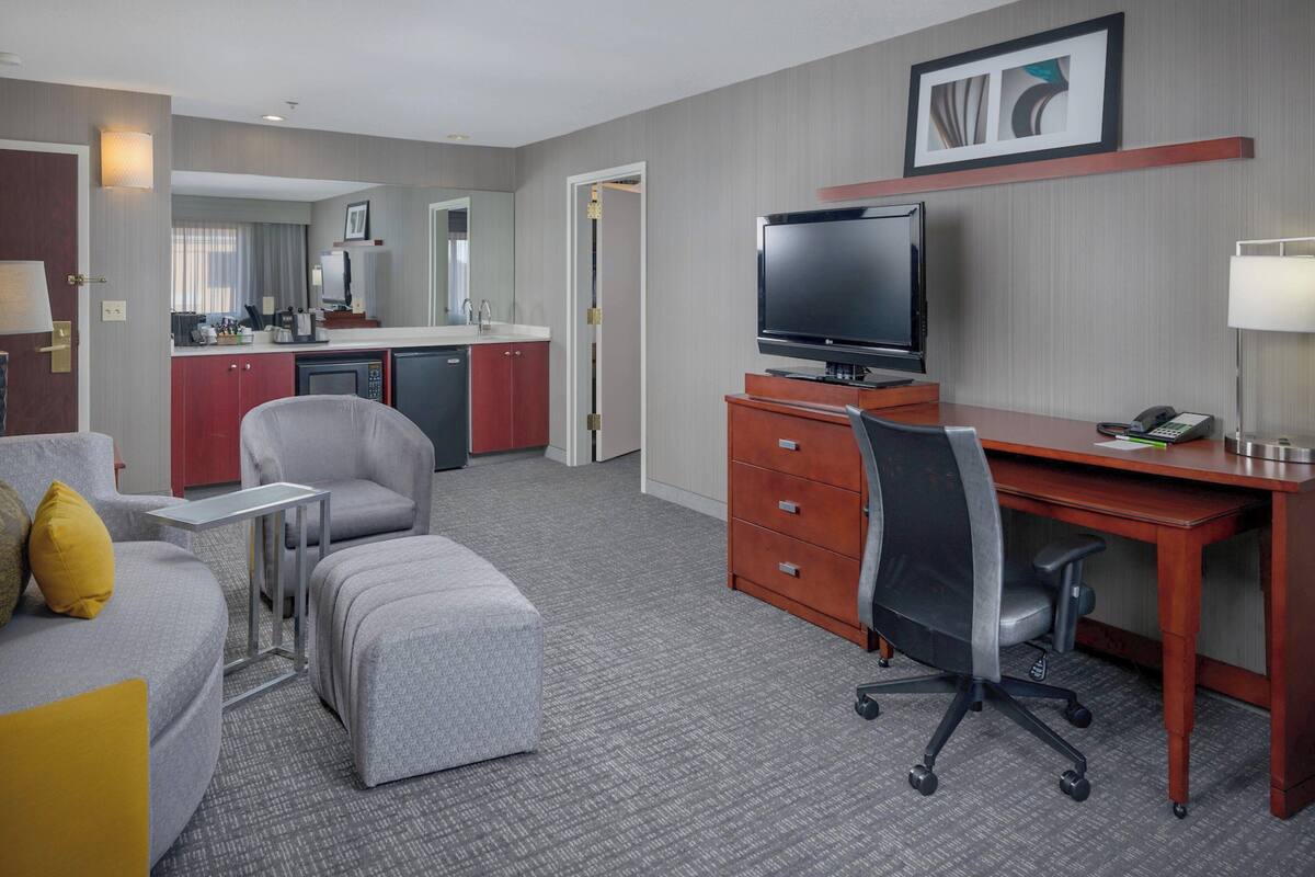 A hotel room with a grey sofa, a small ottoman, a wooden desk with a TV, a chair, a desk lamp, and a kitchenette in the background.