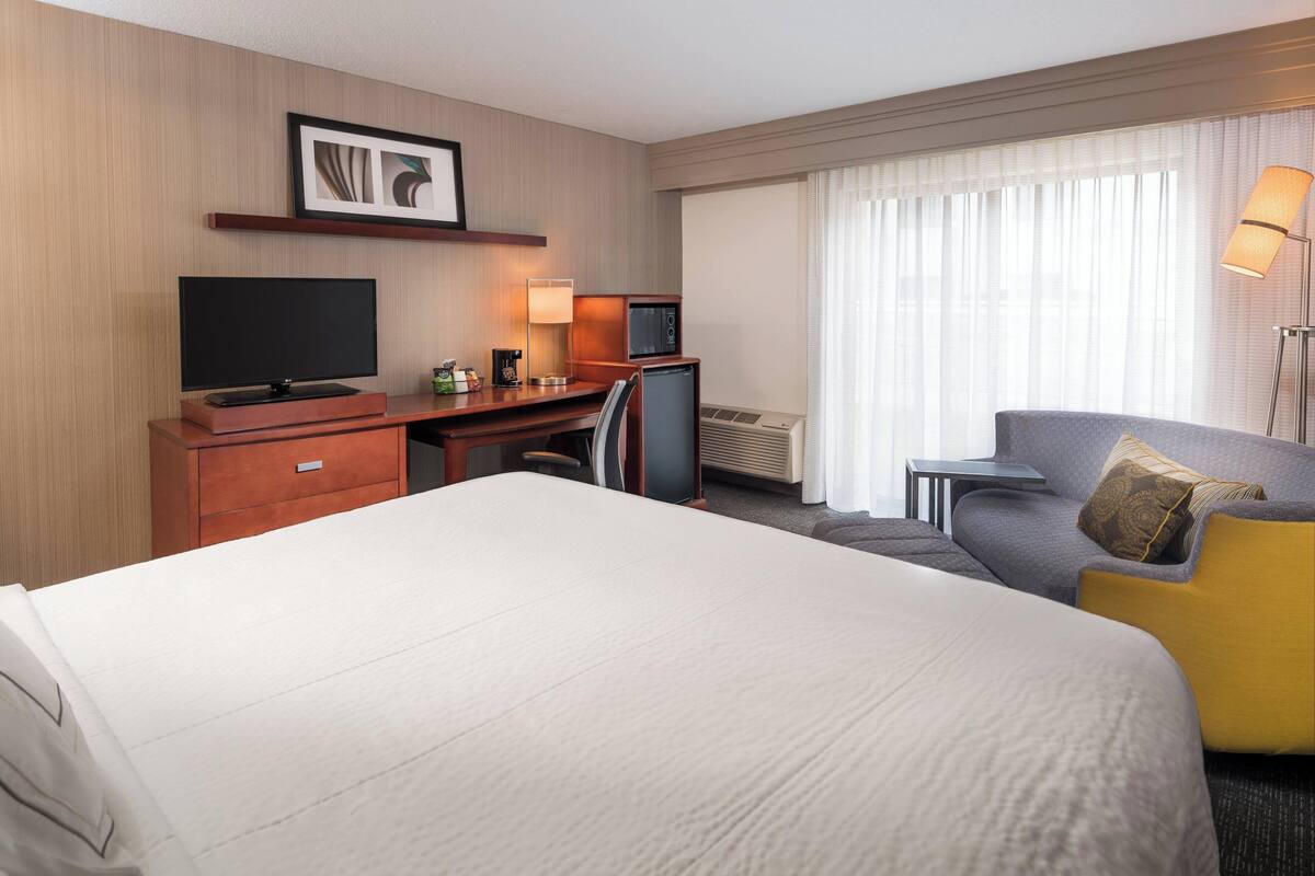 A hotel room with a large bed, a desk with a TV, a small kitchenette, and a sofa with a pillow.
